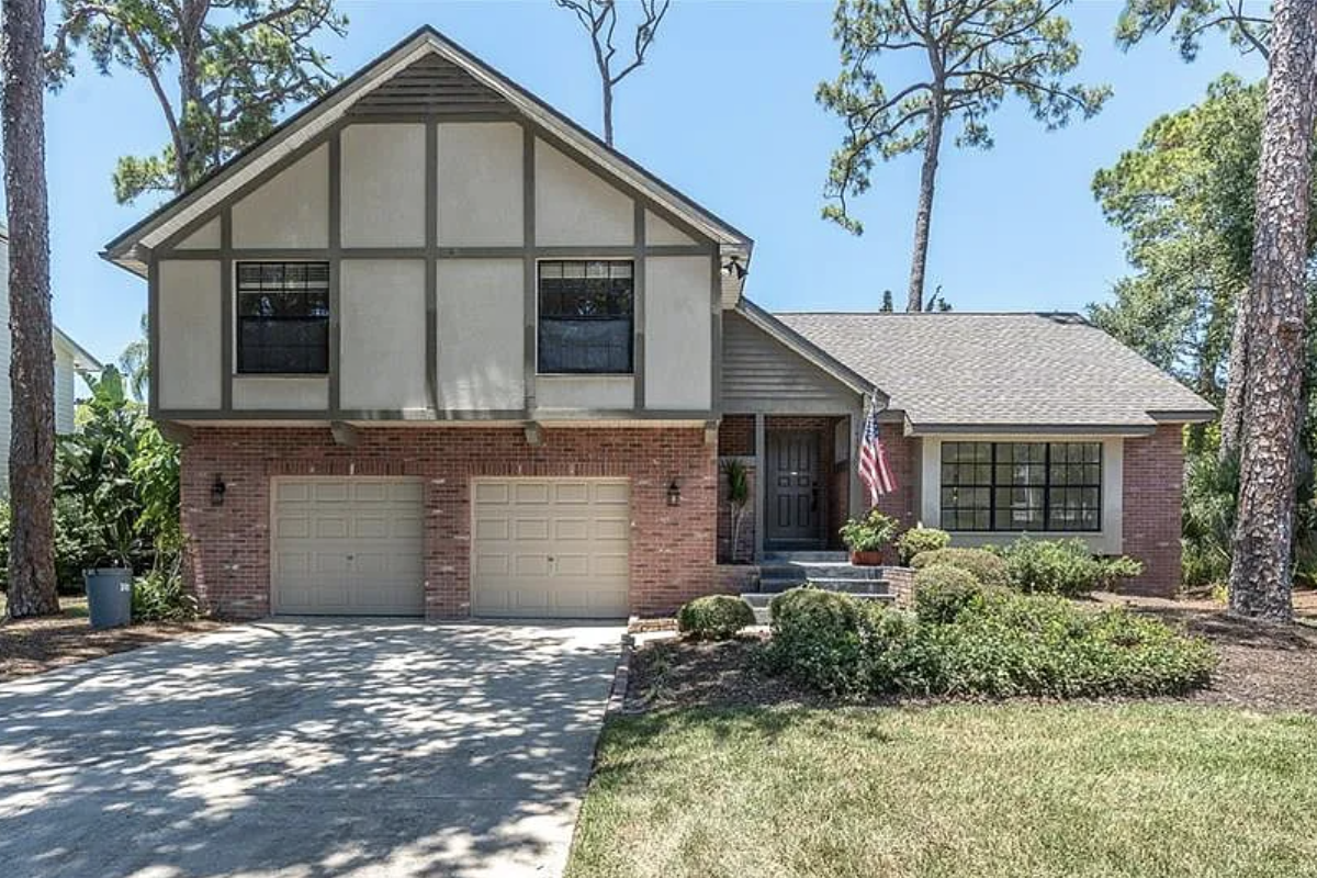 Two-story Tudor-style home with brick lower level and stucco-and-timber upper facade, featuring a two-car garage, covered front entrance with an American flag, landscaped shrubs and lawn, and a long concrete driveway shaded by mature pine trees.